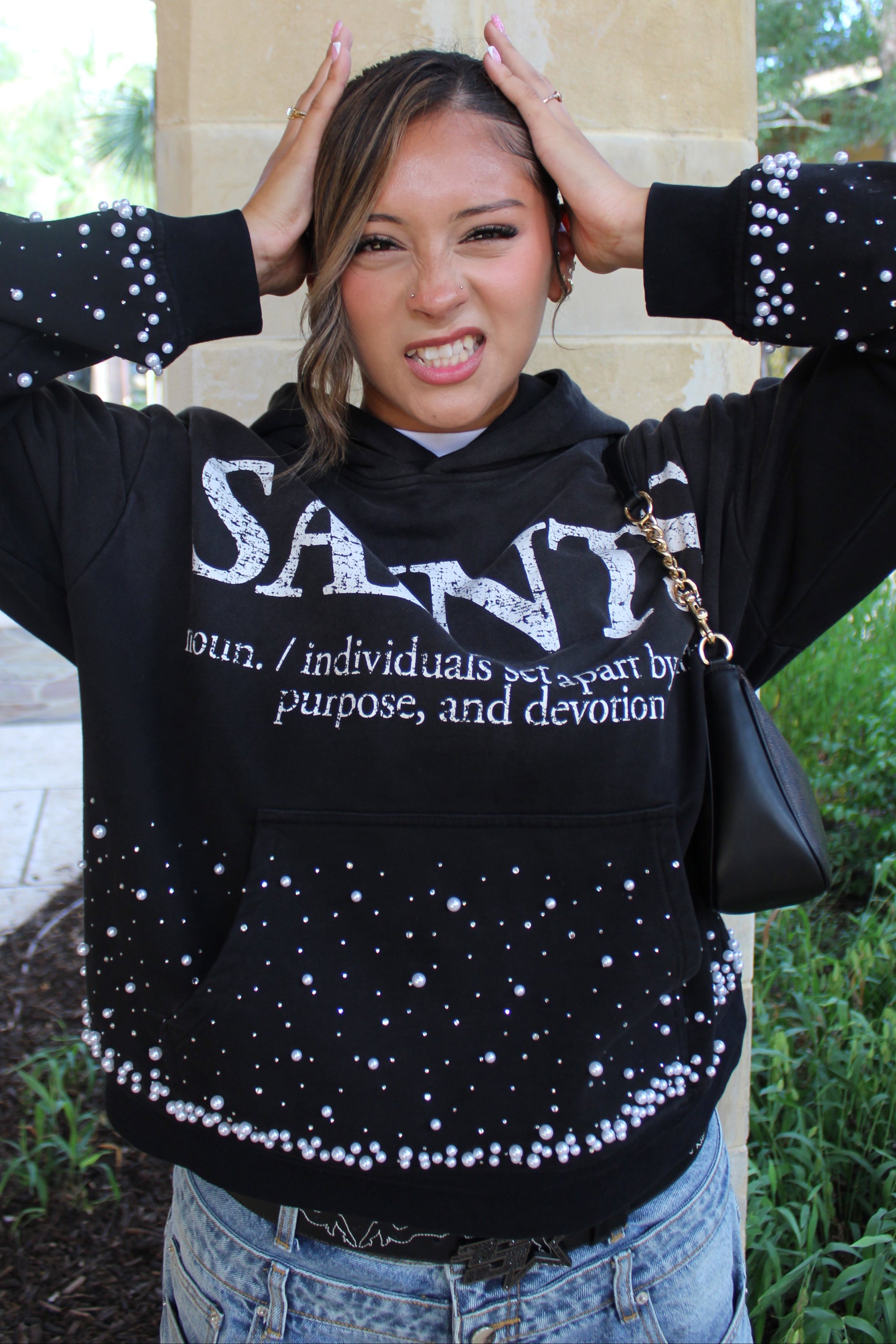 A woman wearing a black long-sleeve sweatshirt with white rhinestone embellishments and a large white 'SAINTS' logo on the front, paired with blue jeans.