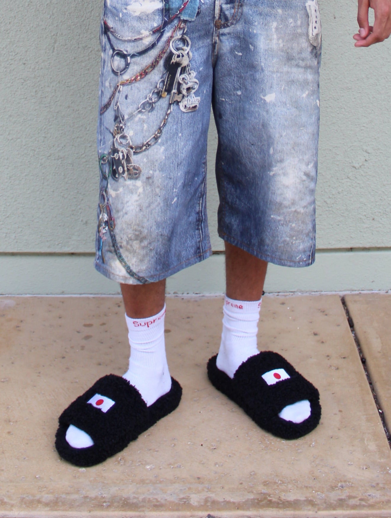 A person stands outdoors against a light gray wall wearing a black hoodie with 'SAINTS' printed in white across the chest, distressed denim shorts, white socks, and black slide sandals.