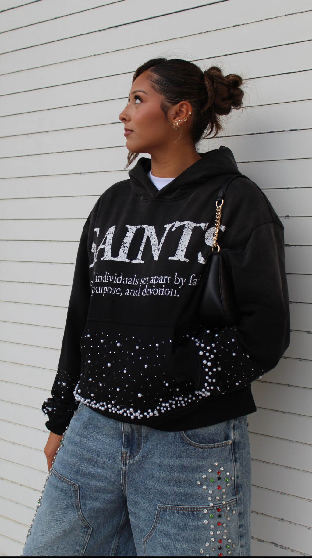 A woman wearing a black Saint Louis Saints hoodie with white lettering and rhinestone embellishments, paired with light blue jeans, standing against a white wood-paneled wall.