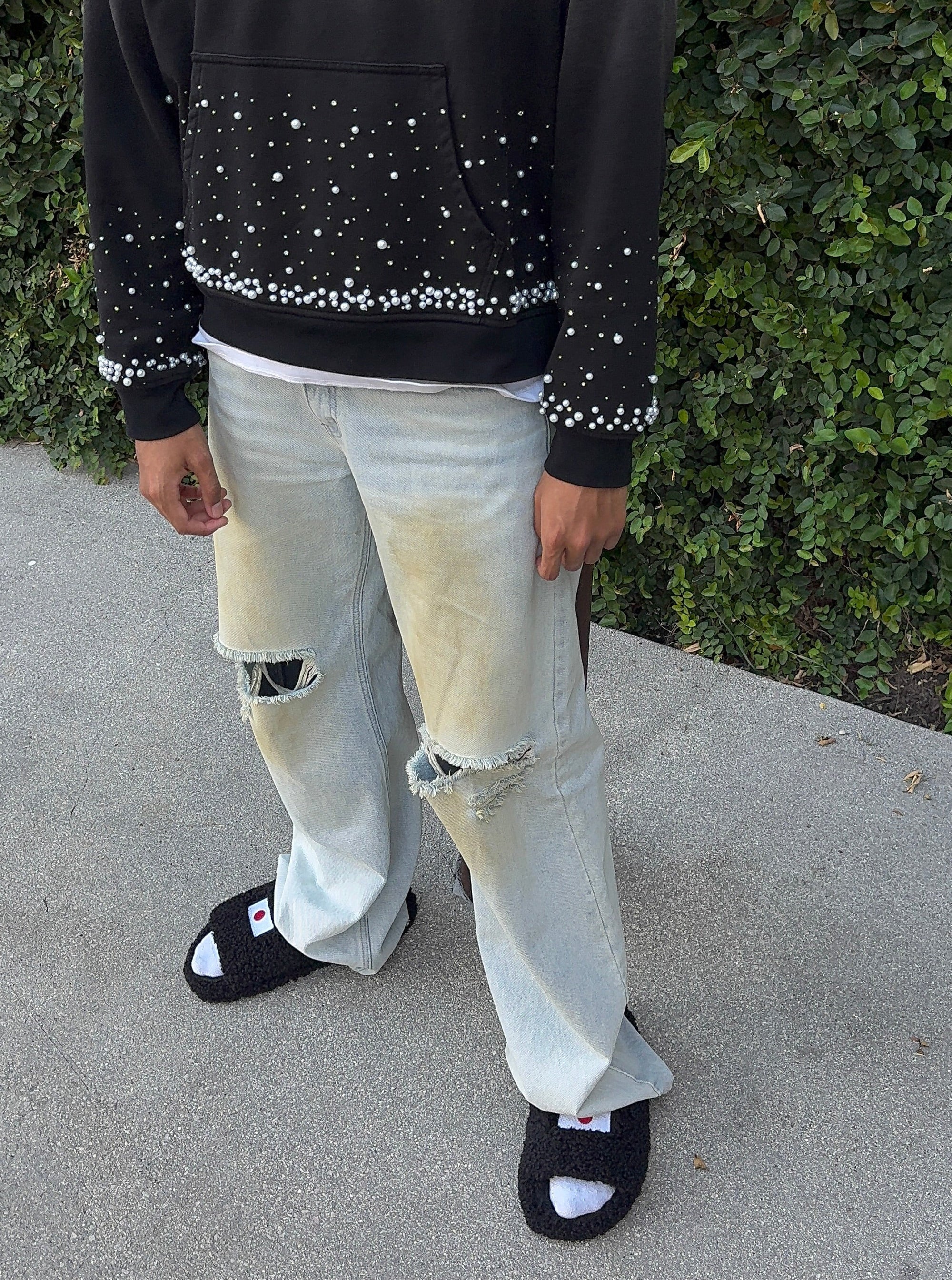 A person wearing a black hoodie with white rhinestone embellishments, light-wash ripped jeans, and black slide sandals, standing outdoors against a hedge and concrete ground.