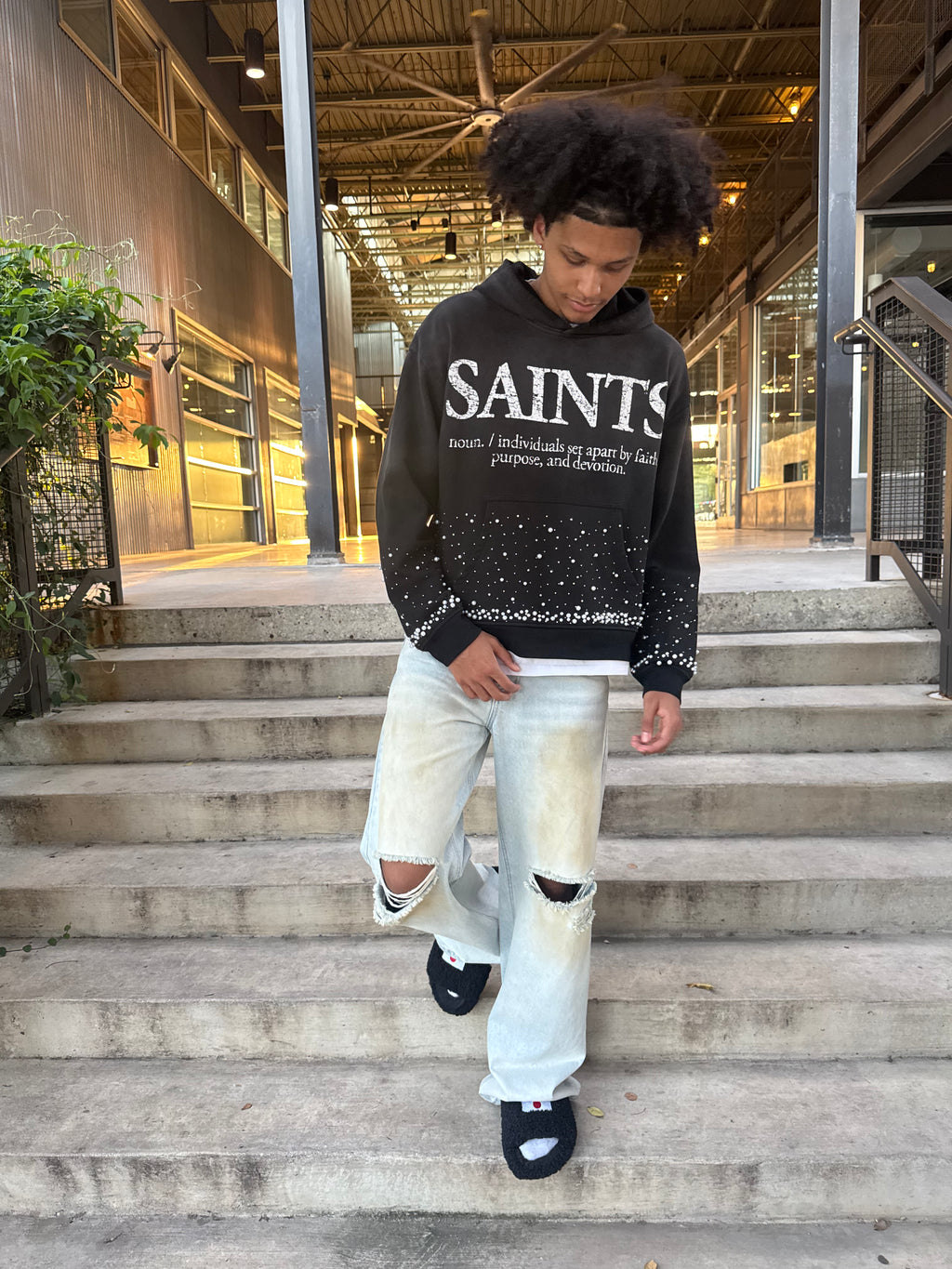 A person standing on concrete stairs wearing a black hoodie with 'SAINTS' printed in white across the chest, light-wash ripped jeans, and black slip-on shoes.