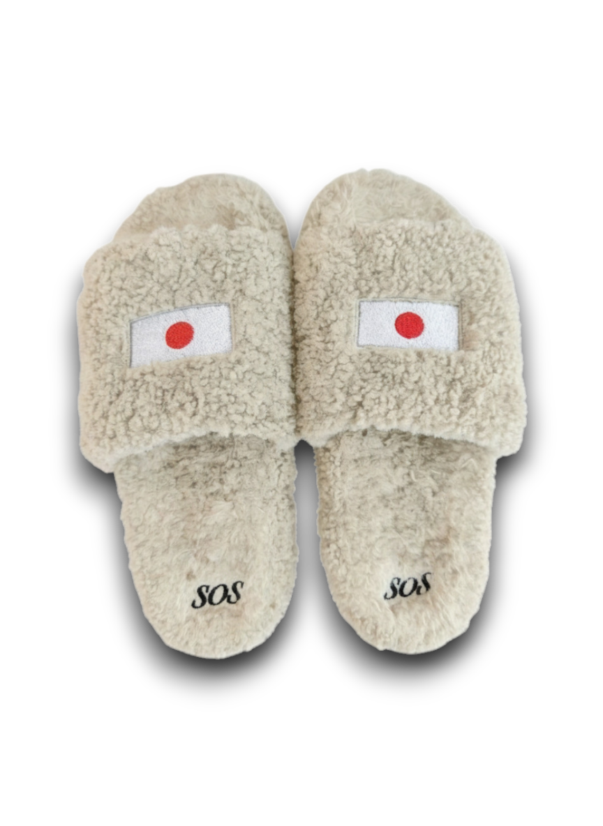 A pair of beige sherpa slide slippers featuring a Japanese flag patch on the top strap and the word 'SOS' embroidered on the sole, set against a plain white background.