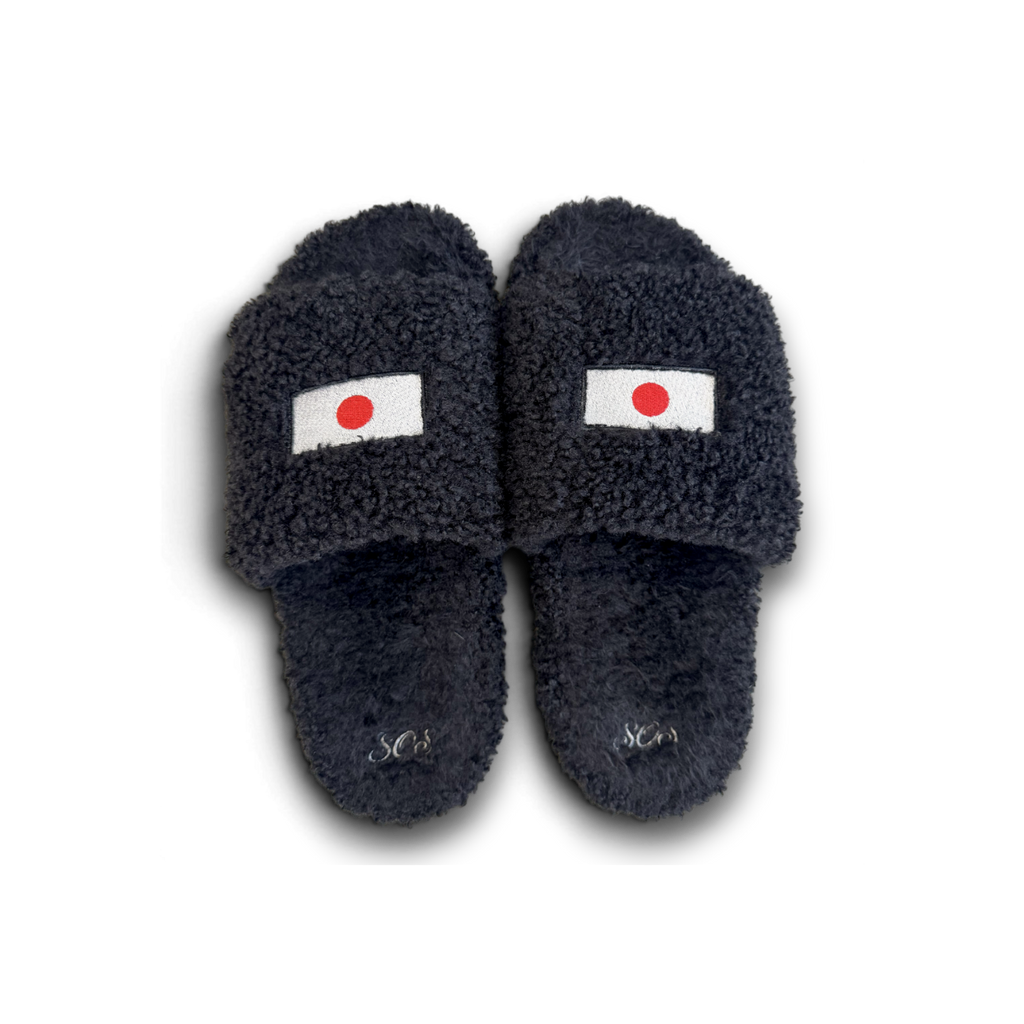A pair of dark navy blue fuzzy slide sandals with a small Japanese flag patch on the top strap and a silver monogram logo on the footbed, shown against a plain white background.