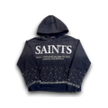 A black hoodie featuring the word 'SAINTS' printed in distressed white letters across the chest, with a dictionary-style definition underneath. The sleeves and lower body of the hoodie are decorated with white rhinestone-like star patterns, and the front pocket is lined with white rhinestones.